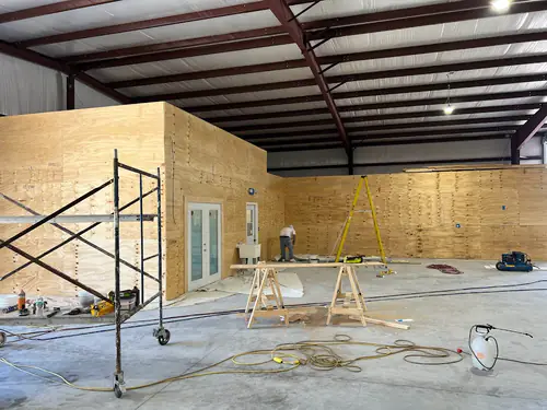 South Daytona industrial park metal building interior before painting, with new unpainted plywood walls, sawhorses, a rolling scaffold and a sprayer
