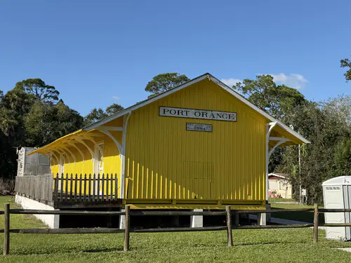 The 1894 Port Orange freight depot at Depot Park after exterior painting, with yellow board and batten siding, white trim and a row of white curved brackets under the platform roof