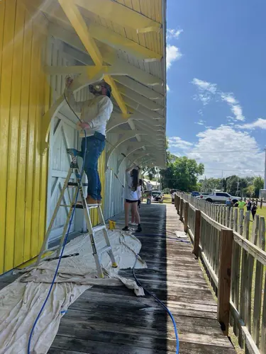Painter on a stepladder spraying the underside of the Port Orange depot platform roof during the job, with drop cloths on the wood deck and an airless spray hose running along it
