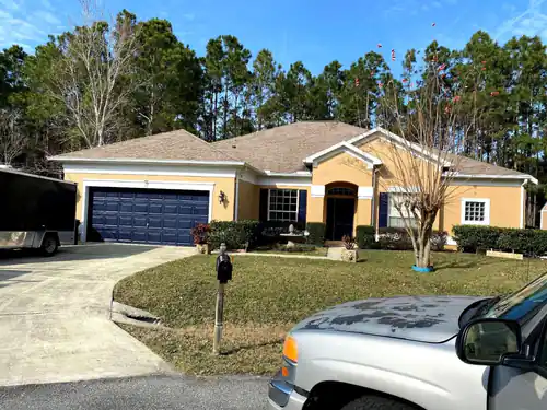 Wider street view of the finished Ormond Beach home with peach stucco walls, white trim and a navy two car garage door