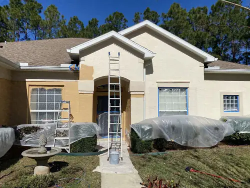 Ormond Beach stucco house during an exterior repaint, with shrubs wrapped in plastic sheeting, windows masked and a ladder standing in the arched entry