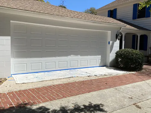 Freshly painted two car raised panel garage door and trim on a pale house, with masking paper still laid over the brick paver driveway