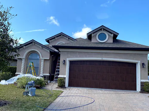 Port Orange stucco house during exterior painting, with an airless paint sprayer on the lawn, plastic over the shrubs and ladders at the arched window