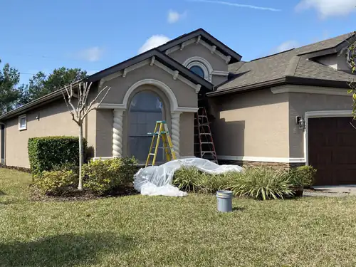 Side of a Port Orange stucco home during painting, with ladders against the gable and the planting bed covered in plastic sheeting