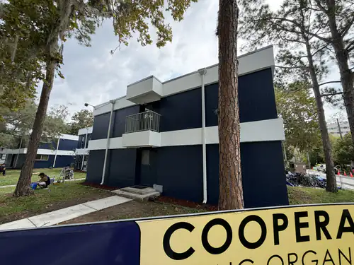 Side of a Gainesville student housing building after painting, navy block with a white band, white balcony and white downspouts among tall pine trees