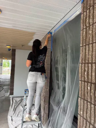 Painter on a stepladder taping the edge of a split face block column in a breezeway during the job, with plastic over the glass and the ceiling half old tan and half new white