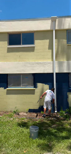 Painter spraying navy onto the lower block wall of a Gainesville student housing building during the job, with the old pale yellow green color still on the rest of the wall and a window masked off